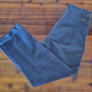 May include: Dark blue denim pants with a straight leg design. The pants have a classic five-pocket style and a Wrangler label on the back pocket. The pants are laid flat on a wooden surface.