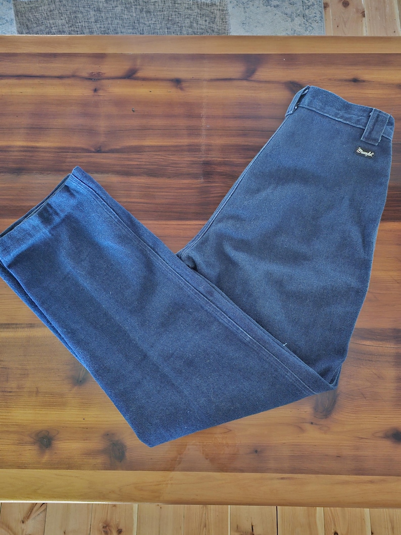 May include: Dark blue denim pants with a straight leg design. The pants have a classic five-pocket style and a Wrangler label on the back pocket. The pants are laid flat on a wooden surface.