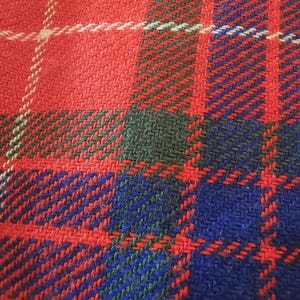 May include: Close-up of a woven fabric with a plaid pattern. The design features a bold combination of red, blue, and green, with white accents. The fabric has a textured appearance, suggesting a warm and durable material.