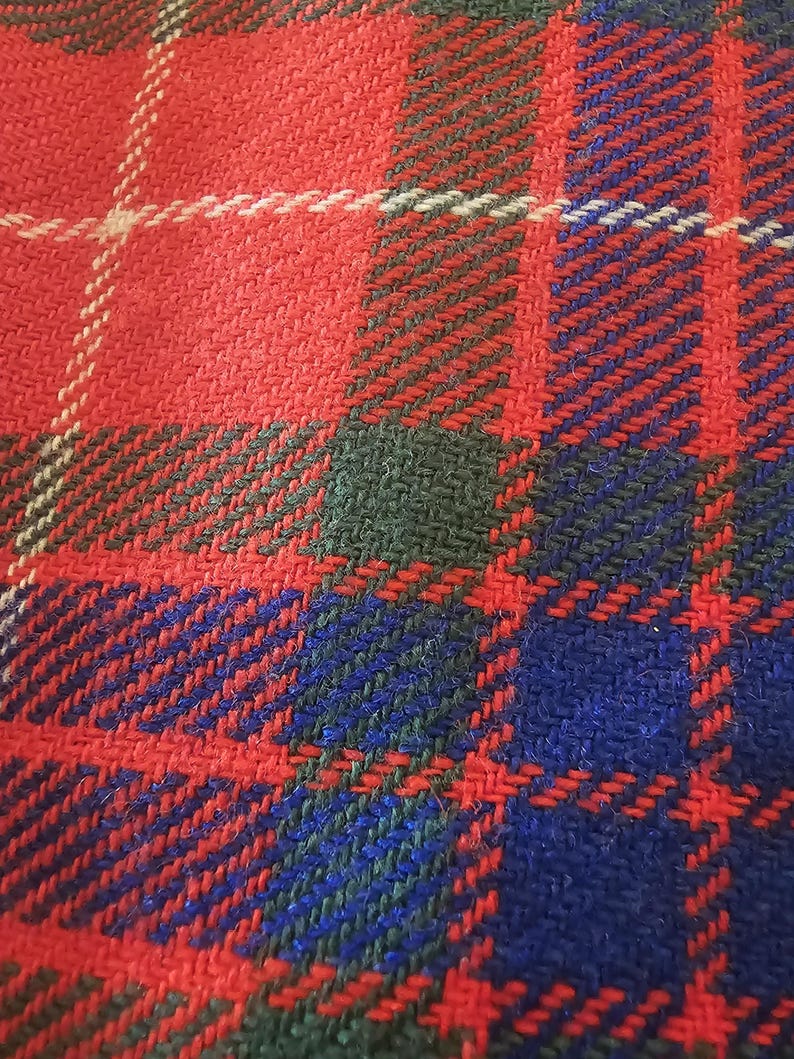 May include: Close-up of a woven fabric with a plaid pattern. The design features a bold combination of red, blue, and green, with white accents. The fabric has a textured appearance, suggesting a warm and durable material.