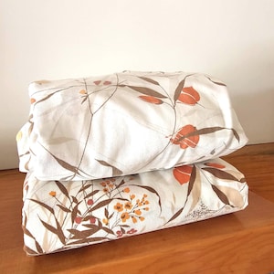 Puede incluir: Dos sábanas blancas dobladas con un estampado floral. El estampado incluye tallos marrones, flores naranjas y pequeños detalles amarillos y rojos. Las sábanas están apiladas una encima de la otra.