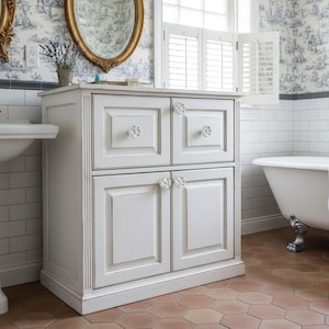 May include: A white bathroom cabinet with four doors and decorative floral knobs. The cabinet is in a bathroom with a white pedestal sink, a claw-foot bathtub, and blue and white wallpaper.