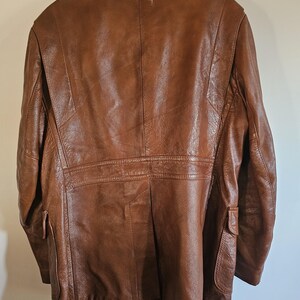 May include: A brown leather jacket with a classic design. The jacket features a notched collar, two front pockets, and a tailored fit. The leather has a rich, textured appearance, and the jacket is hanging on a white hanger.