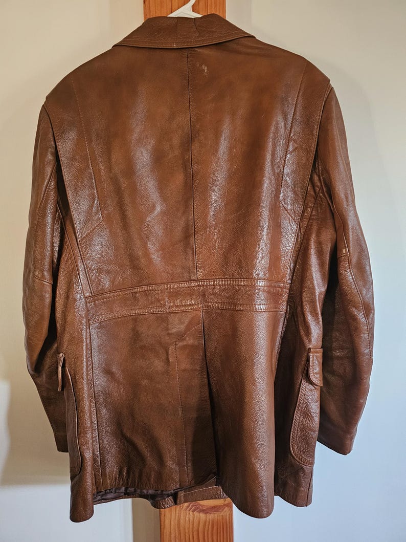 May include: A brown leather jacket with a classic design. The jacket features a notched collar, two front pockets, and a tailored fit. The leather has a rich, textured appearance, and the jacket is hanging on a white hanger.