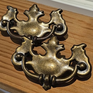 May include: Two antique brass drawer pulls with ornate, leaf-like designs. Each pull features a curved handle and decorative backplate. The hardware has a vintage aesthetic, perfect for furniture restoration projects.