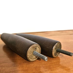 May include: Two dark brown wooden furniture legs with brass-coloured metal ends and threaded bolts. The legs are cylindrical and appear to be new. The background is a light wood surface.