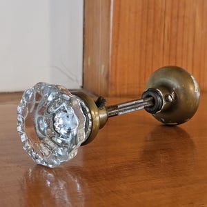 May include: A vintage door knob set featuring a clear, faceted glass knob on one end and a smooth, tarnished brass-toned metal knob on the other. The two knobs are connected by a central metal spindle with a visible set screw and mounting hardware.