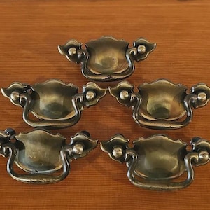 May include: Five antique brass drawer pulls with a curved handle and decorative floral accents. The pulls have a tarnished, aged appearance, suggesting they are vintage or vintage-inspired. The hardware is arranged on a wooden surface.