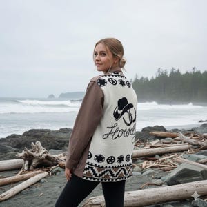 May include: A brown and cream-coloured jacket with a cowboy hat and the word "Howdy" embroidered on the back. The jacket has brown sleeves and black and white geometric patterns. The person is standing on a beach.