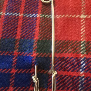 May include: A large, shiny silver safety pin. The pin is set against a backdrop of a red, blue, and green plaid fabric. The pin's design includes a looped end and a clasp.
