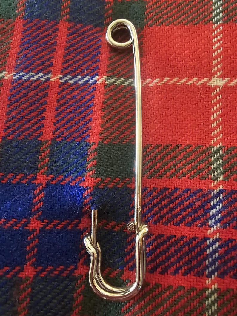 May include: A large, shiny silver safety pin. The pin is set against a backdrop of a red, blue, and green plaid fabric. The pin's design includes a looped end and a clasp.