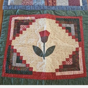 May include: A two-part quilted textile piece featuring a top section with a multi-coloured patchwork grid of red, blue, and white fabrics, and a bottom section with a central tulip appliqué surrounded by a log cabin pattern in red and dark tones.