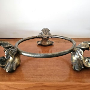 May include: An antique brass-toned metal ring holder with decorative leaf and scroll details. The ring is supported by three decorative elements, with a circular ring in the centre. The holder is designed to sit on a flat surface.