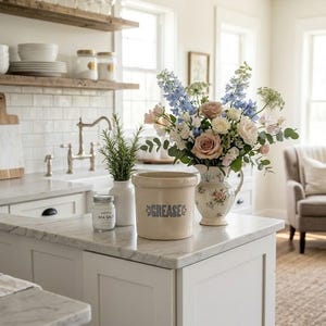 May include: A kitchen island with a marble worktop displays a floral arrangement in a decorative jug. A beige crock with "GREASE" printed on it and a jar of sea salt are also on the counter. The kitchen has white cabinets and shelves.
