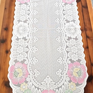 May include: Oval white lace table runner with a floral design. The runner features pink, yellow, and green flowers at each end, with a detailed, openwork pattern in the center. The runner is placed on a wooden table.