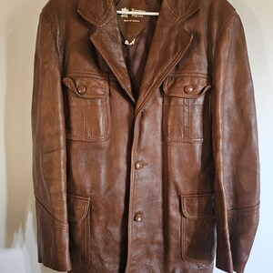 May include: A brown leather jacket with a classic design. The jacket features a notched collar, four front pockets, and button closures. The jacket is hanging on a white hanger.