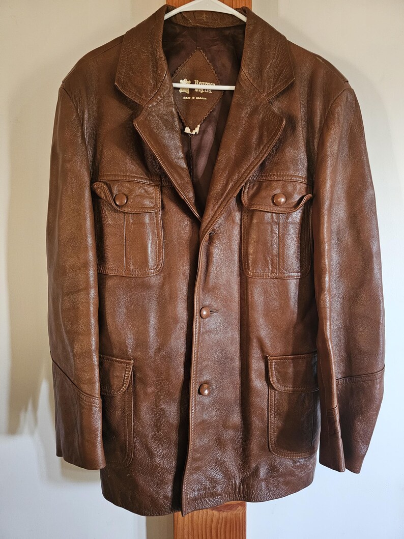 May include: A brown leather jacket with a classic design. The jacket features a notched collar, four front pockets, and button closures. The jacket is hanging on a white hanger.