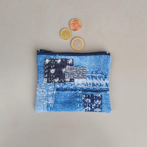 Op de afbeelding: Een rechthoekig denim etui met ritssluiting, voorzien van een patchworkontwerp met diverse blauwe stofresten, witte handgestikte details en drie metalen munten die boven het etui zijn geplaatst. Het etui heeft een donkerblauwe rits.