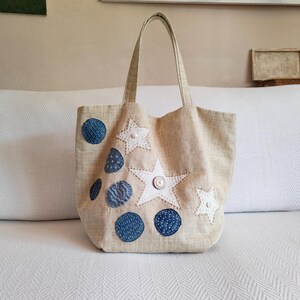 Può includere: Borsa tote beige con applicazioni a forma di stelle bianche e cerchi blu. La borsa ha due manici e una chiusura con bottone. La borsa è realizzata in tessuto color naturale.