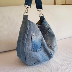 Könnte beinhalten: Hobo-Tasche aus hellblauem Jeansstoff. Die Tasche hat eine Tasche auf der Vorderseite und einen Jeansriemen mit silberfarbenen Metallbeschlägen. Das Innenfutter ist gemustert. Die Tasche liegt auf einer weißen Oberfläche.