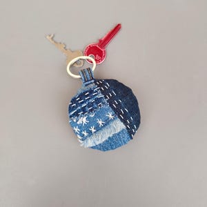 以下が含まれることがあります： 白いステッチと星が付いた丸い青いデニムのキーホルダー。キーホルダーはリサイクルデニム生地で作られています
