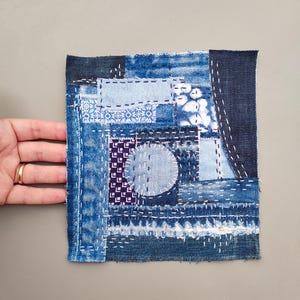 May include: A square textile patch featuring a patchwork design of various blue denim fabrics and patterned cotton scraps. The pieces are joined with visible white running stitch embroidery, including a central circular motif and frayed edges.