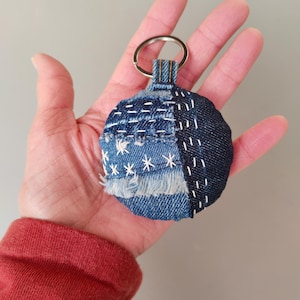 Puede incluir: Un llavero de mezclilla azul con forma redonda y un anillo de llave plateado. El llavero está hecho de diferentes piezas de mezclilla cosidas juntas con hilo blanco.