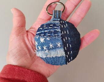 May include: A blue denim keychain with a round shape and a silver key ring. The keychain is made of different pieces of denim sewn together with white thread.