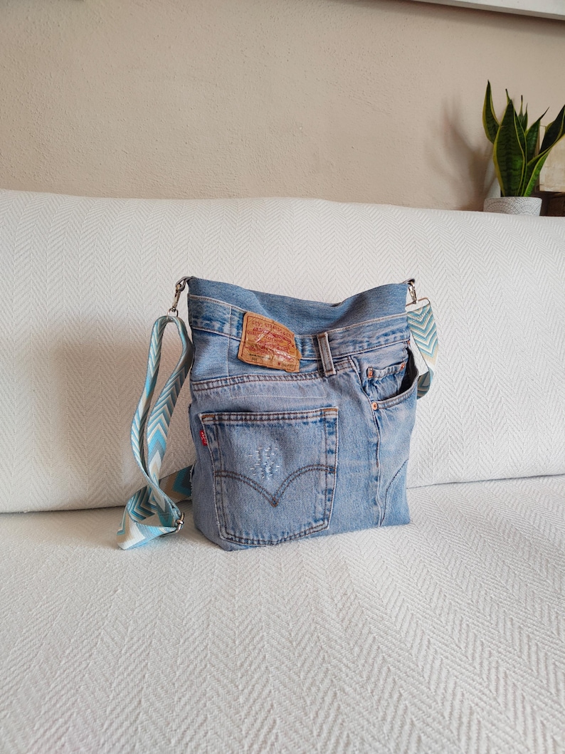 a faded blue jeans crossbody bag with jacquard multicolor strap, the jeans pockets and the original jeans tag.
The adjustable strap has a geometric design in light blue, beige, dusty blue and white colors. The two swivel hooks are silver tone.
Bag standing on a white sofa.