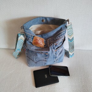 a faded blue denim bag slightly open showing the beige interior lining with one pocket.
The bag is standing on a white sofa, in front of the bag there is one black cell phone and one black e-reader.