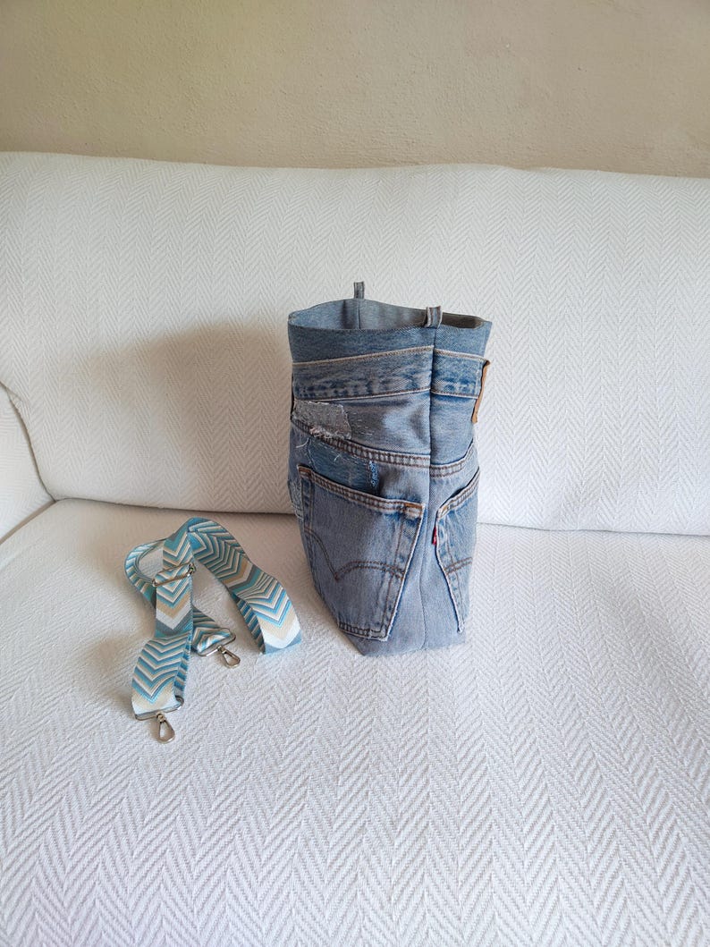 Puede incluir: Un bolso tote de mezclilla hecho de jeans reciclados, con una correa estampada en azul claro y blanco. El bolso tiene un dise&ntilde;o cl&aacute;sico de cinco bolsillos, con costuras visibles y una etiqueta roja de Levi's. El bolso mide aproximadamente 30 cm de alto.