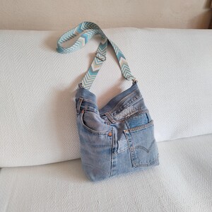a faded blue jeans crossbody bag with geometric jacquard strap leaning against the back of a white sofa.
The adjustable strap has a geometroc design in light blue, beige, dusty blue and white colors.
Hardware in silver tone.