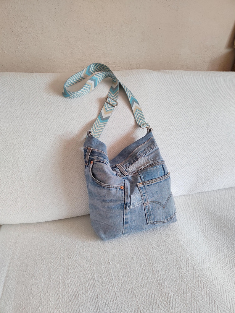 a faded blue jeans crossbody bag with geometric jacquard strap leaning against the back of a white sofa.
The adjustable strap has a geometroc design in light blue, beige, dusty blue and white colors.
Hardware in silver tone.