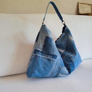 May include: A denim patchwork tote bag with a single shoulder strap. The bag is made from various shades of blue denim, with visible stitching and a unique, handcrafted appearance. The bag is sitting on a white surface.