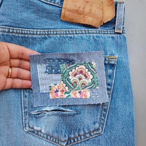 Może przedstawiać: Zbliżenie jasnoniebieskich dżinsów z naszytą kieszenią. Łatka to prostokąt materiału z kwiatowym wzorem w kolorze różowym, zielonym i pomarańczowym. Dżinsy mają postarzany wygląd z podartą kieszenią.