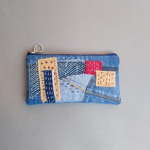 May include: A rectangular denim pouch with a zipper closure and a small metal ring. The pouch is adorned with patches of various fabrics in shades of blue, beige, and red, stitched together in a patchwork design. The overall aesthetic is handcrafted and unique.