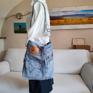 Puede incluir: Bolso cruzado de mezclilla azul claro hecho de jeans reciclados. El bolso presenta los bolsillos traseros originales, un parche de cuero rojo y marr&oacute;n y una correa estampada. El bolso se exhibe en un maniqu&iacute; con una camisa blanca.