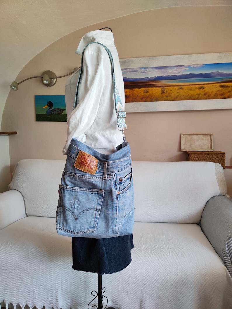 Puede incluir: Bolso cruzado de mezclilla azul claro hecho de jeans reciclados. El bolso presenta los bolsillos traseros originales, un parche de cuero rojo y marr&oacute;n y una correa estampada. El bolso se exhibe en un maniqu&iacute; con una camisa blanca.