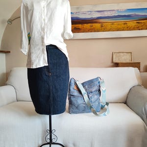 a light blue denim bag with multicolor adjustable strap standing on a white sofa, at its side there is a black iron mannequin wearing a white shirt and a dark blue denim skirt.
The jacquard polyester strap has a geometric design in light blue, beige, dusty blue and off white colors.