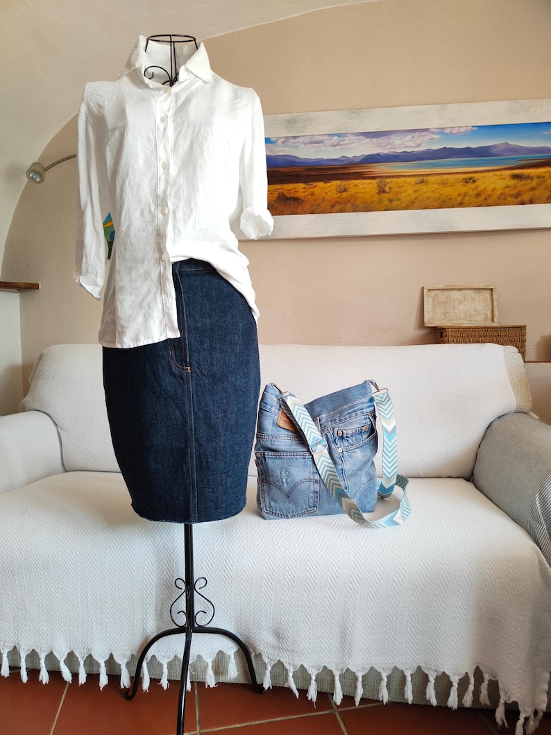 a light blue denim bag with multicolor adjustable strap standing on a white sofa, at its side there is a black iron mannequin wearing a white shirt and a dark blue denim skirt.
The jacquard polyester strap has a geometric design in light blue, beige, dusty blue and off white colors.