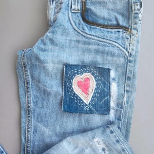 May include: Light blue denim jeans with a heart-shaped patch. The patch features a pink heart with white stitching. The jeans have a classic five-pocket design and a faded wash, with some distressed details.