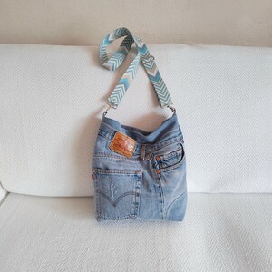 a crossbody bag handmade from upcycled faded blue jeans, visible mending hand stitched, original jeans tag and geometric jacquard strap.
The bag is standing on a white sofa.