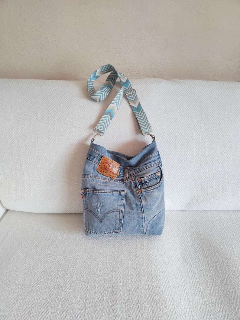 a crossbody bag handmade from upcycled faded blue jeans, visible mending hand stitched, original jeans tag and geometric jacquard strap.
The bag is standing on a white sofa.