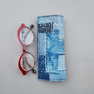 May include: A pair of red-rimmed glasses with tortoiseshell temples positioned next to a rectangular denim case. The case is constructed from various shades of blue denim fabric patches joined together with visible white hand-stitched embroidery.