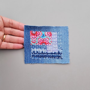 May include: A small square denim patch with a colourful floral fabric patch sewn on top. The denim patch has visible stitching and frayed edges.