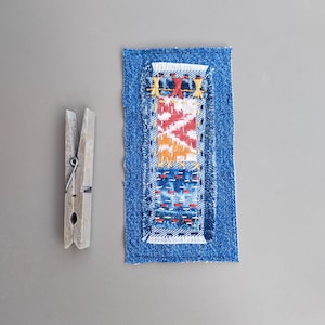 May include: A small, rectangular denim patch with a colorful embroidered design. The embroidery features red, orange, and blue patterns. A wooden clothespin is positioned to the left of the patch. The background is a solid gray.