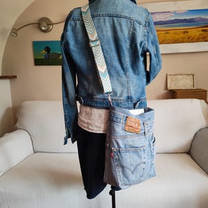 a faded blue denim bag worn across an iron mannequin with a denim jacket , a white shirt and a dark blue denim skirt on.
The mannequin is on its back side.