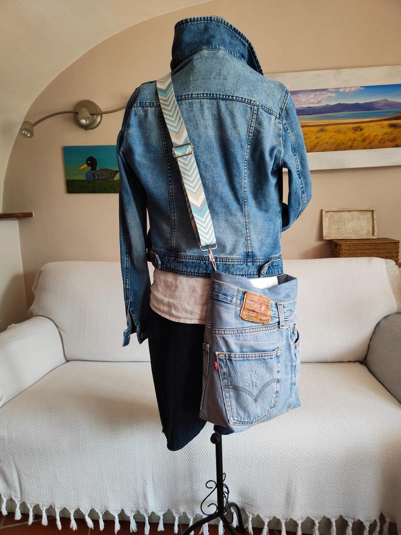 a faded blue denim bag worn across an iron mannequin with a denim jacket , a white shirt and a dark blue denim skirt on.
The mannequin is on its back side.