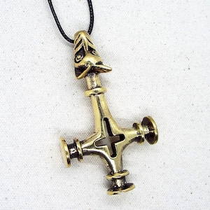 May include: A gold-coloured pendant in the shape of a cross with a bird's head at the top. The cross has a hole in the centre and is attached to a black cord.