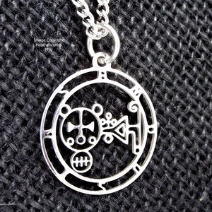 May include: Silver pendant necklace with a circular design featuring a detailed, intricate symbol. The symbol is surrounded by a circle with a border.